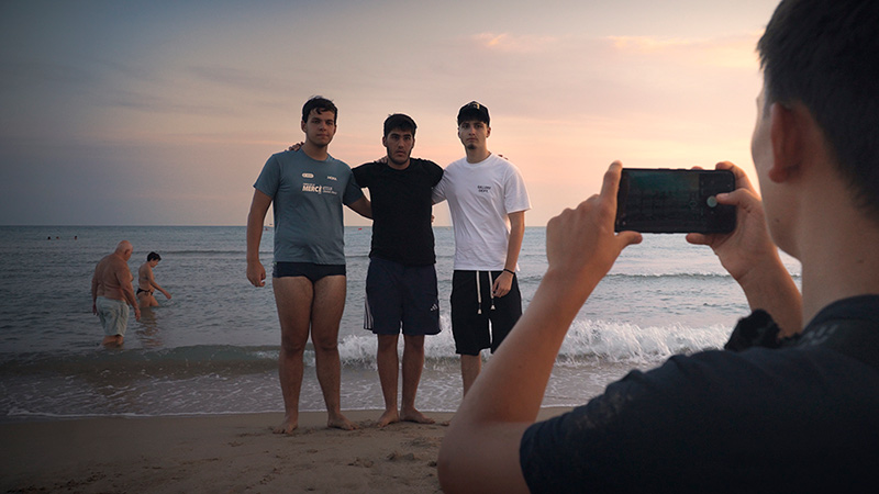Fotograma de DUES CARES: tres amics posen a la platja al capvespre mentre un altre els fa una foto amb el mòbil