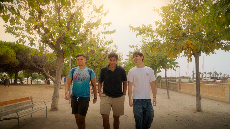 Fotograma de DUES CARES: tres amics passegen i riuen per un passeig amb arbres a prop del port